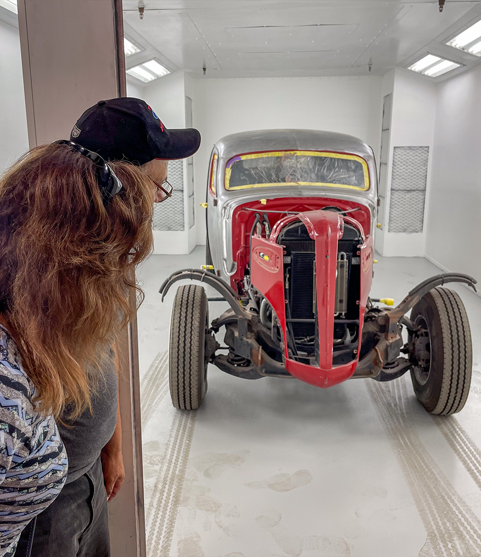 A couple peeking into the paint booth to look at a hot rod in for paint